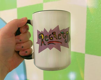 May include: White ceramic mug with a black handle. The mug has a purple, yellow, green, and blue graphic that says "Regrets" in a stylized font.