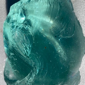 May include: A large, translucent, teal-colored piece of sea glass with a swirling pattern. The glass has a smooth, polished surface and is free of any visible cracks or chips.