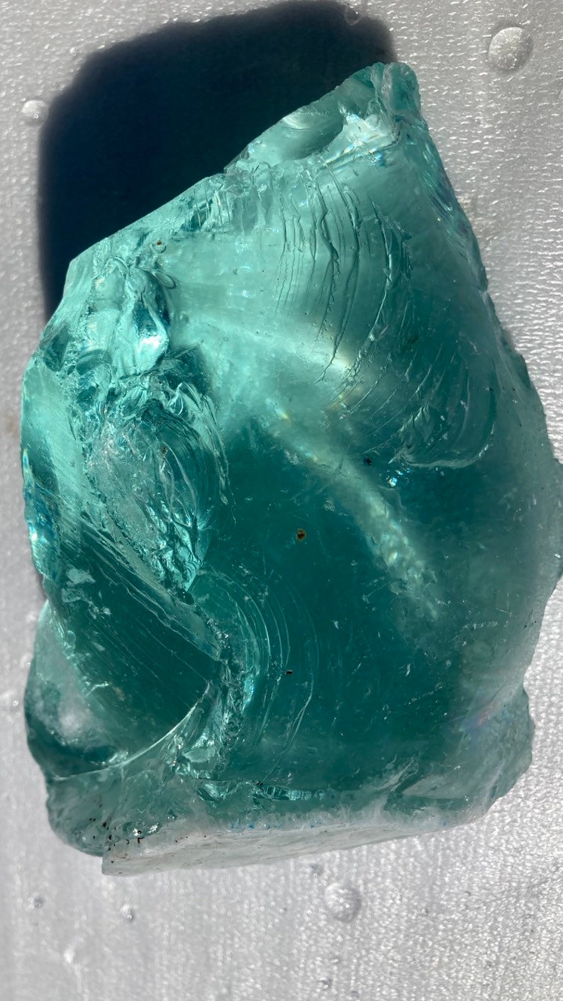 May include: A large, translucent, teal-colored piece of sea glass with a swirling pattern. The glass has a smooth, polished surface and is free of any visible cracks or chips.