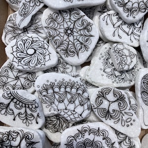 May include: A collection of white painted rocks with black ink mandala designs. The rocks are various shapes and sizes and are arranged in a pile.