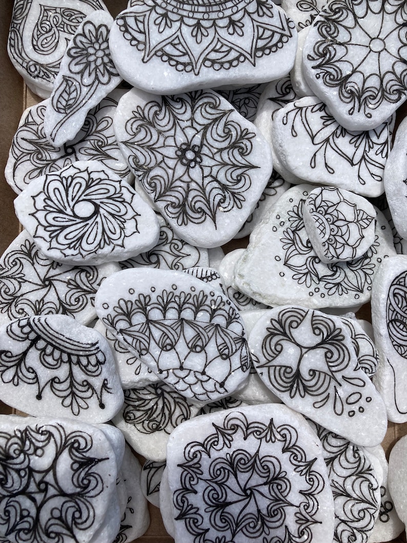 May include: A collection of white painted rocks with black ink mandala designs. The rocks are various shapes and sizes and are arranged in a pile.