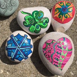 May include: Five painted rocks with different designs. One rock has a green clover design, one has a blue snowflake design, one has a pink heart design, one has a red and orange geometric design, and one has a black and white swirl design.