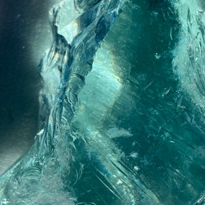 May include: A large, clear, teal-colored crystal with a rough, textured surface. The crystal is illuminated by a light source, creating a bright, reflective effect.