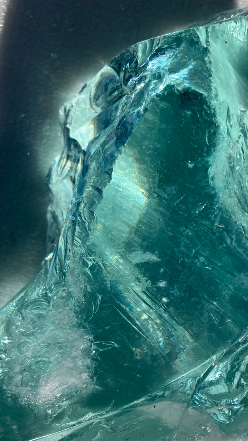 May include: A large, clear, teal-colored crystal with a rough, textured surface. The crystal is illuminated by a light source, creating a bright, reflective effect.