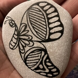 May include: A gray stone painted with a black and white butterfly design. The butterfly has intricate details and a floral pattern.