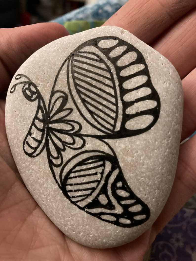 May include: A gray stone painted with a black and white butterfly design. The butterfly has intricate details and a floral pattern.