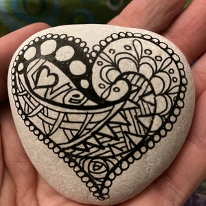 May include: A white heart-shaped rock painted with black ink. The heart is filled with intricate patterns and swirls.