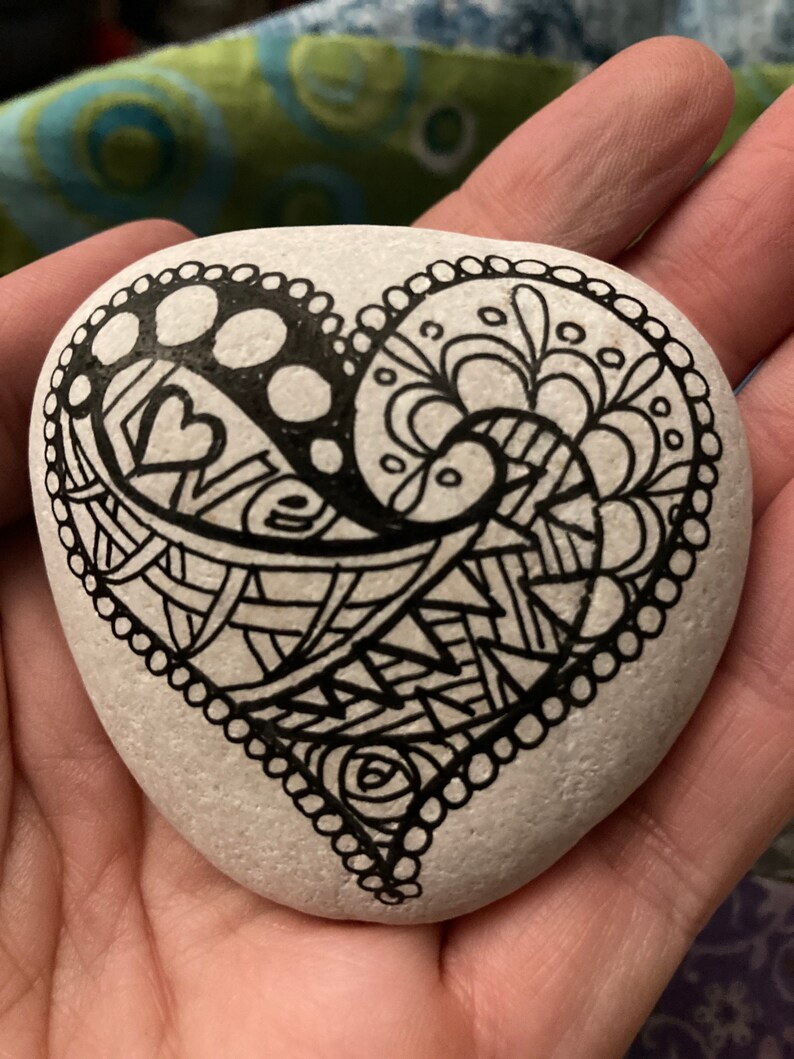 May include: A white heart-shaped rock painted with black ink. The heart is filled with intricate patterns and swirls.