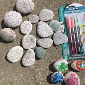 May include: A collection of painted rocks with intricate designs, including mandalas, hearts, and flowers. The rocks are arranged on a sandy surface. A pack of gel pens is also included in the image. The pens are labeled "Gel Pens" and "Stylos de Gel".