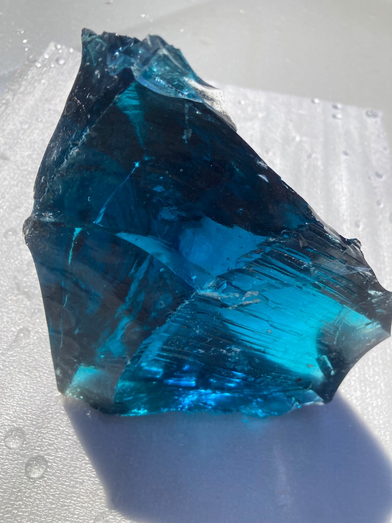 May include: A large, teal blue piece of sea glass with a smooth, polished surface. The glass has a slightly cloudy appearance, giving it a unique and natural look.