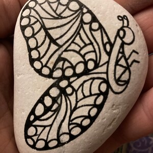 May include: A white rock painted with a black and white butterfly design. The butterfly has intricate details and a swirling pattern.