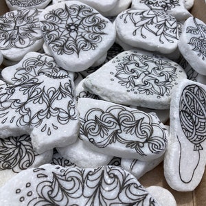 May include: A collection of white stones with black ink drawings of various designs, including swirls, flowers, and geometric patterns.