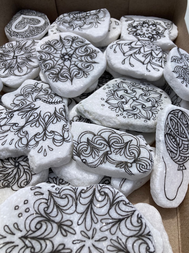 May include: A collection of white stones with black ink drawings of various designs, including swirls, flowers, and geometric patterns.