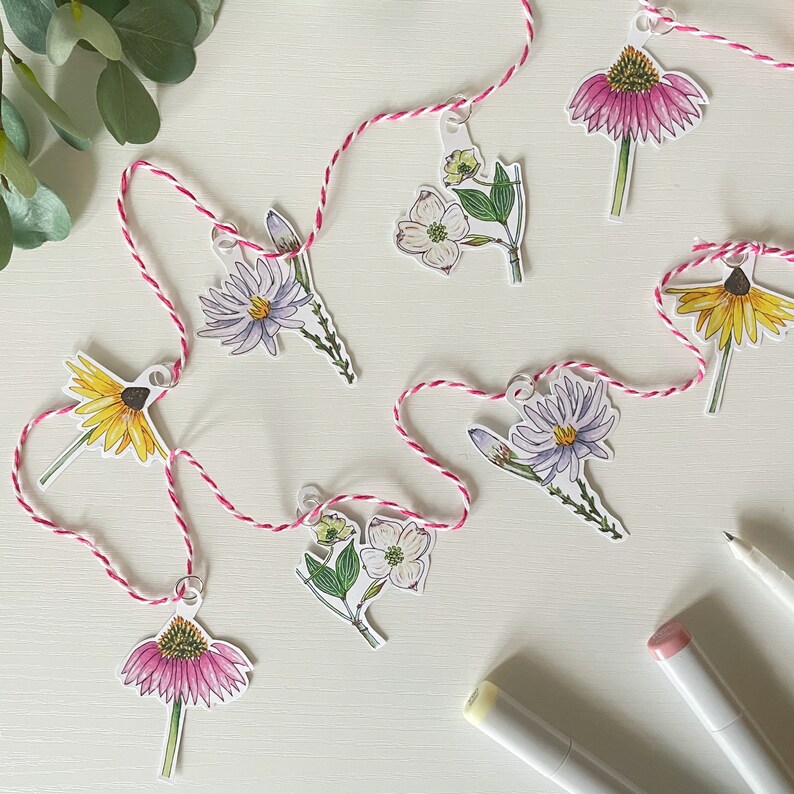 May include: A floral garland featuring various flower illustrations, including purple, yellow, and pink blooms. The paper cutouts are strung together with a pink and white twine. The garland is set against a white background, with art supplies visible.