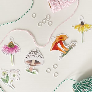 May include: A collection of botanical-themed paper cutouts, including a pink coneflower, a yellow daisy, and an orange mushroom. Silver jump rings and pink and green twine are scattered around the cutouts, suggesting a crafting project.