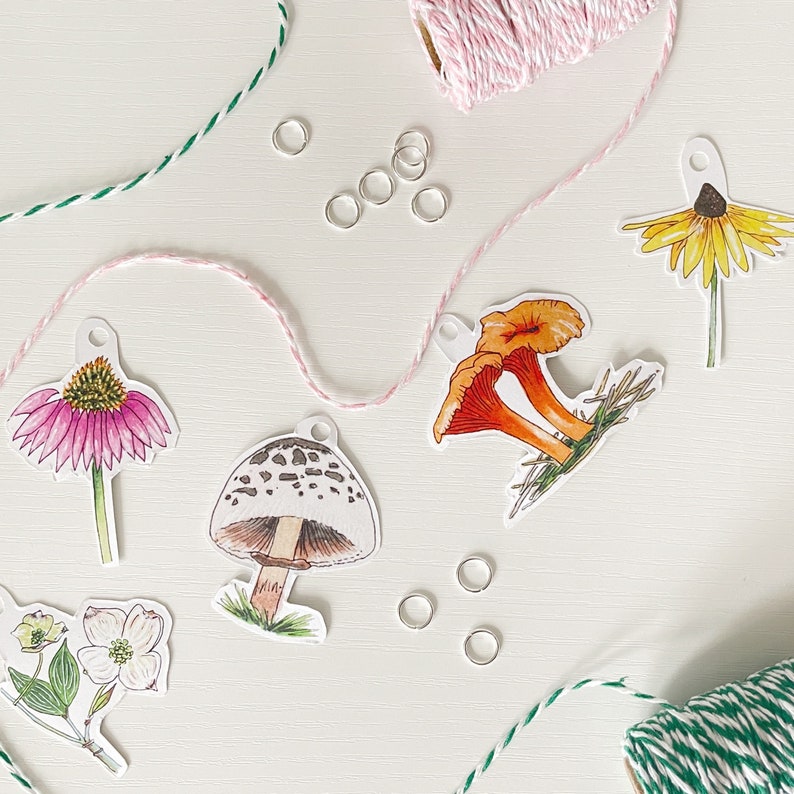 May include: A collection of botanical-themed paper cutouts, including a pink coneflower, a yellow daisy, and an orange mushroom. Silver jump rings and pink and green twine are scattered around the cutouts, suggesting a crafting project.