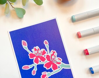 May include: A vibrant floral illustration on a blue background. The artwork showcases pink flowers with white and light green accents. Several grey markers with coloured tips are visible, along with a small bowl and a sprig of greenery.