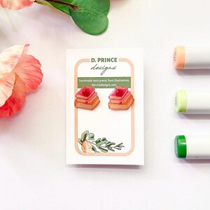 Peut inclure: Une paire de boucles d'oreilles en forme de piles de livres dans des tons rose et orange, présentées sur une carte blanche D. Prince Designs avec une illustration florale, le texte D. PRINCE designs et handmade resin pieces from illustrations.