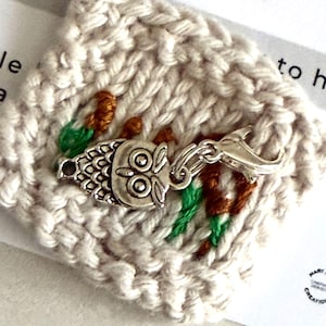 May include: A small, cream-coloured knitted square with embroidered brown and green details and a silver owl charm attached. The charm has a lobster clasp. Text on the card reads "A little hug just for you."