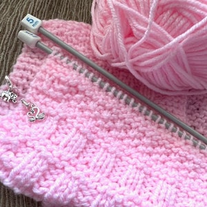 May include: Pink yarn and knitting needles with a pink knitted fabric. The needles are 5 mm and there are two stitch markers in the shape of a swan and an elephant.