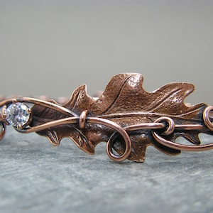 May include: A copper wire bracelet with a detailed oak leaf design and a single clear rhinestone.