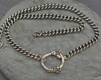 May include: A silver-toned necklace with a curb chain design. The necklace features a circular pendant with a textured, organic design. The clasp is a hook and eye style. The jewelry is displayed on a gray stone surface.