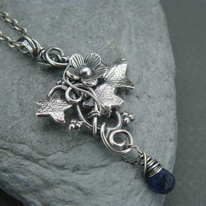 Peut inclure: Collier en argent avec un pendentif représentant une délicate fleur et des feuilles de lierre, accentué d'une pierre précieuse bleu foncé.