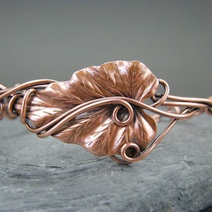 Puede incluir: Un brazalete de alambre de cobre con un gran diseño de hoja de cobre texturizado. La hoja está centrada en el brazalete y está rodeada de un delicado trabajo de alambre.