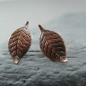 May include: A pair of copper leaf earrings with a textured surface. The earrings are a light brown color and have a delicate, organic shape.