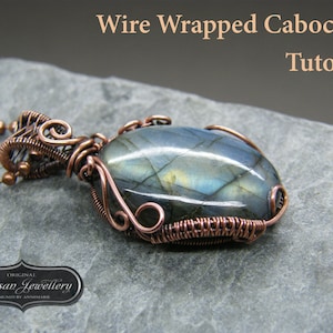 Peut inclure: Un pendentif en fil de cuivre enveloppé avec une pierre cabochon grise et bleue. Le pendentif est attaché à une chaîne argentée. L'image comprend le texte "Original Artisan Jewellery Designed by Annemarie".