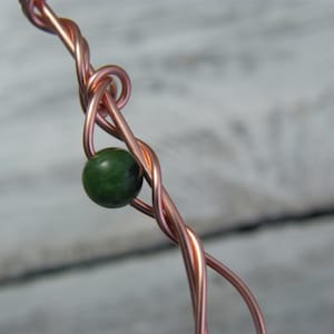 May include: A close-up of a copper wire wrapped around a green gemstone bead. The wire is twisted and forms a knot around the bead.
