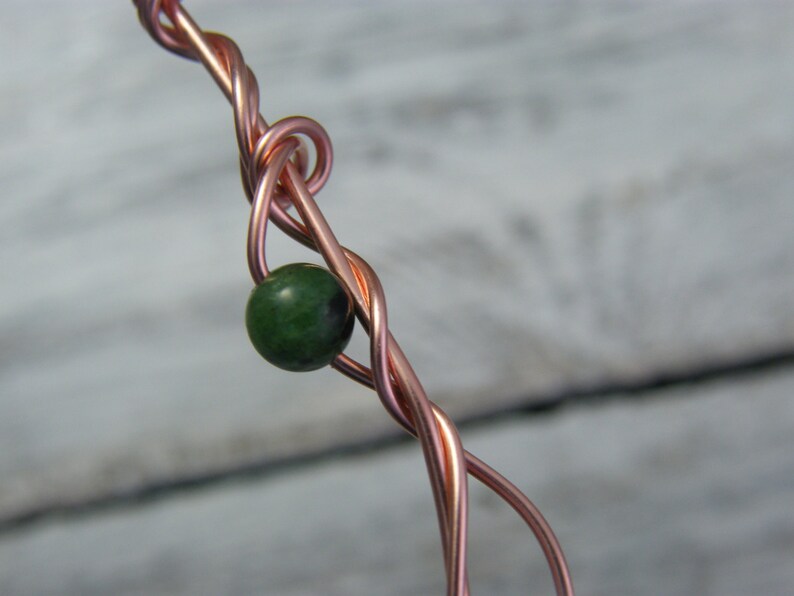 May include: A close-up of a copper wire wrapped around a green gemstone bead. The wire is twisted and forms a knot around the bead.