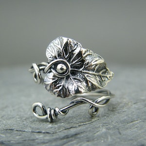 Puede incluir: Un anillo de plata con un diseño de hoja. La hoja es detallada y texturizada, y el anillo está hecho de alambre que está retorcido y envuelto alrededor de la hoja.
