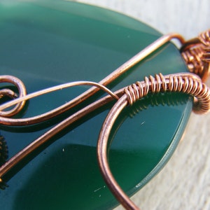 May include: Close-up of a green gemstone pendant with copper wire wrapping. The wire is wrapped around the stone in a decorative pattern.