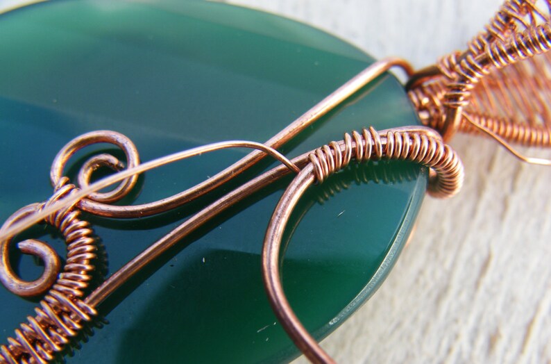 May include: Close-up of a green gemstone pendant with copper wire wrapping. The wire is wrapped around the stone in a decorative pattern.