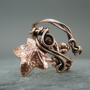 Könnte beinhalten: Ein Kupferring mit einem gehämmerten Efeublatt-Design und einem roten Edelstein. Der Ring ist aus Draht gefertigt und hat ein filigranes, kompliziertes Design.