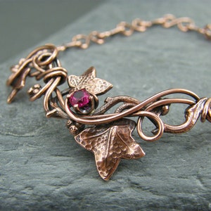 Op de afbeelding: Een koperen armband met een delicaat klimopontwerp en een roze edelsteen. De bladeren zijn getextureerd en de ranken zijn gedraaid.
