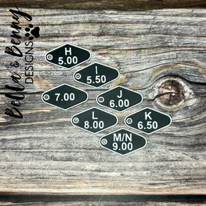 May include: Black and white metal tags with letters and numbers. The tags are in a variety of sizes and are arranged in a row on a wooden surface. The tags are labeled with the letters H, I, J, K, L, and M/N, and the numbers 5.00, 5.50, 6.00, 6.50, 7.00, 8.00, and 9.00.
