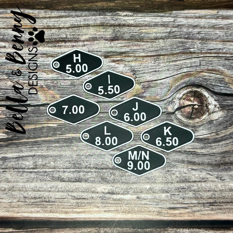 May include: Black and white metal tags with letters and numbers. The tags are in a variety of sizes and are arranged in a row on a wooden surface. The tags are labeled with the letters H, I, J, K, L, and M/N, and the numbers 5.00, 5.50, 6.00, 6.50, 7.00, 8.00, and 9.00.