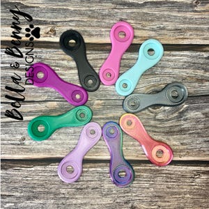 May include: A collection of colorful, oval-shaped accessories in various colors, including pink, purple, green, and blue. Each piece has a circular hole at each end and a textured surface. The items are arranged on a wooden surface.