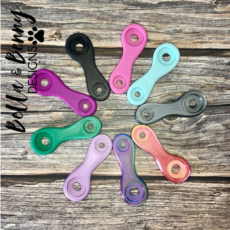 May include: A collection of colorful, oval-shaped accessories in various colors, including pink, purple, green, and blue. Each piece has a circular hole at each end and a textured surface. The items are arranged on a wooden surface.