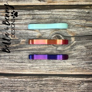 May include: Three colorful, curved, bar-shaped objects in shades of mint green, iridescent orange-red, and purple-blue are arranged vertically on a wooden surface. The text "Bella & Benny DESIGNS" is printed in black script on the left side.