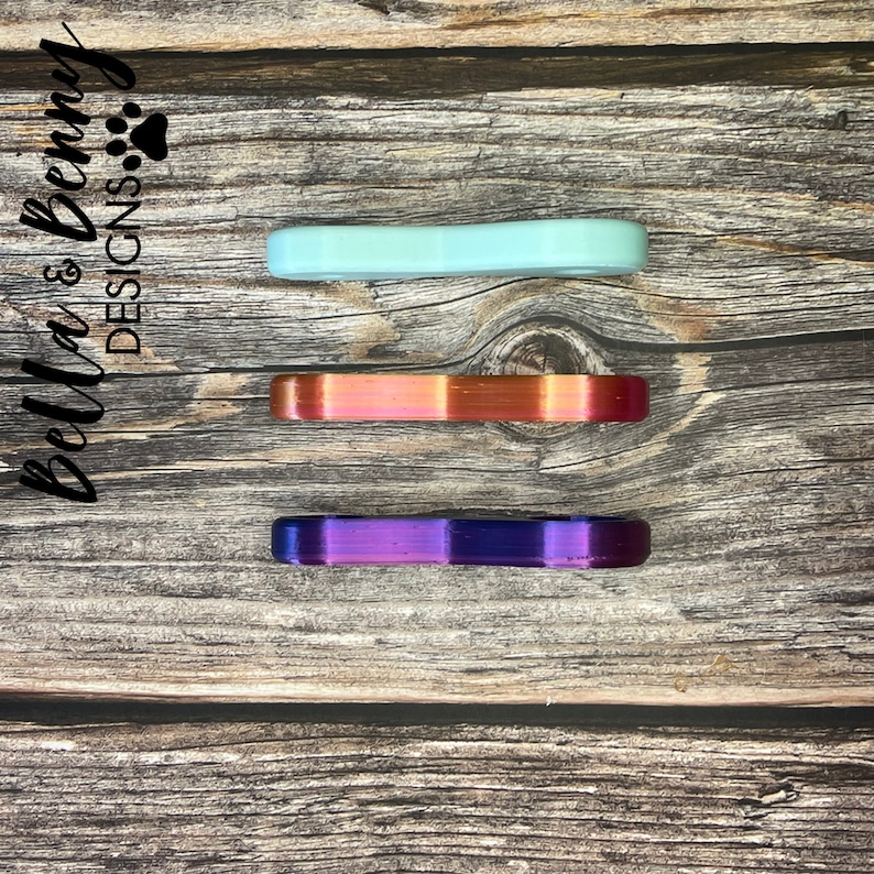 May include: Three colorful, curved, bar-shaped objects in shades of mint green, iridescent orange-red, and purple-blue are arranged vertically on a wooden surface. The text "Bella & Benny DESIGNS" is printed in black script on the left side.