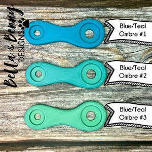 May include: Three blue and teal ombre-colored, dumbbell-shaped objects with circular holes. Each object has a different shade of blue and teal. The text "Blue/Teal Ombre #1", "Blue/Teal Ombre #2", and "Blue/Teal Ombre #3" is displayed.