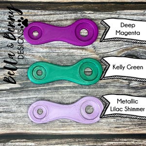 May include: Three plastic keychains in different colors: deep magenta, kelly green, and metallic lilac shimmer. Each keychain has a unique shape with two holes.