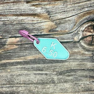May include: A turquoise blue key tag with a white border and white text that reads "K 6.50". The key tag is attached to a pink plastic key ring.