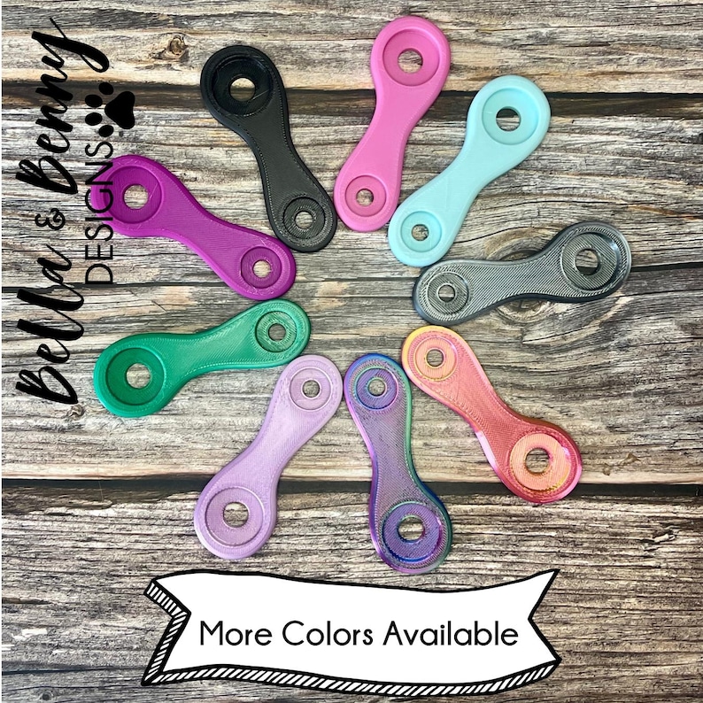 May include: A collection of colorful, uniquely shaped accessories in various hues, including pink, green, and purple. The accessories have a distinctive design with a circular opening at each end and a connecting bar. The image includes the text "More Colors Available."