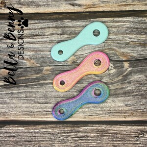 May include: Three colorful, small, oval-shaped objects with a hole at each end. The top one is light blue, the middle one is a mix of pink and yellow, and the bottom one is a rainbow of colors. The objects are on a wooden surface.