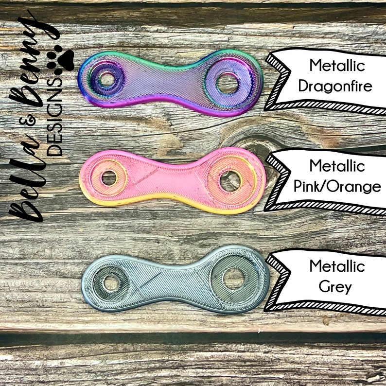 May include: Three metallic fidget toys in Dragonfire, Pink/Orange, and Grey. Each has a unique color gradient and a chain link design with two circular openings. The toys are displayed on a rustic wooden surface.
