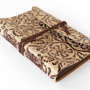 May include: A beige leather journal with a floral embossed design. The journal is secured with a brown braided leather cord. The cover features a flap closure. The edges of the pages are visible.